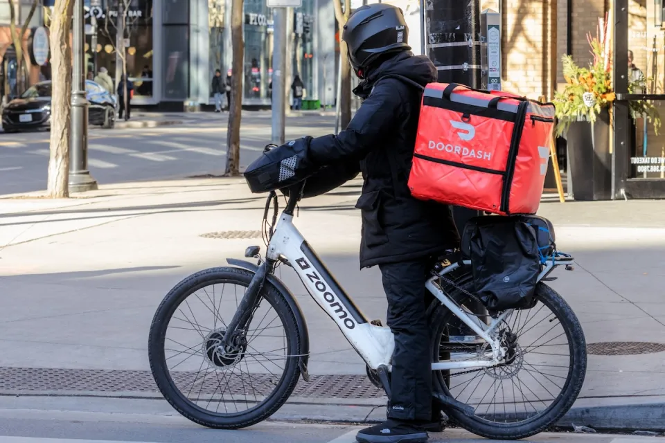 Person in black clothing and helmet with red DoorDash box on back of electric bike on urban street.